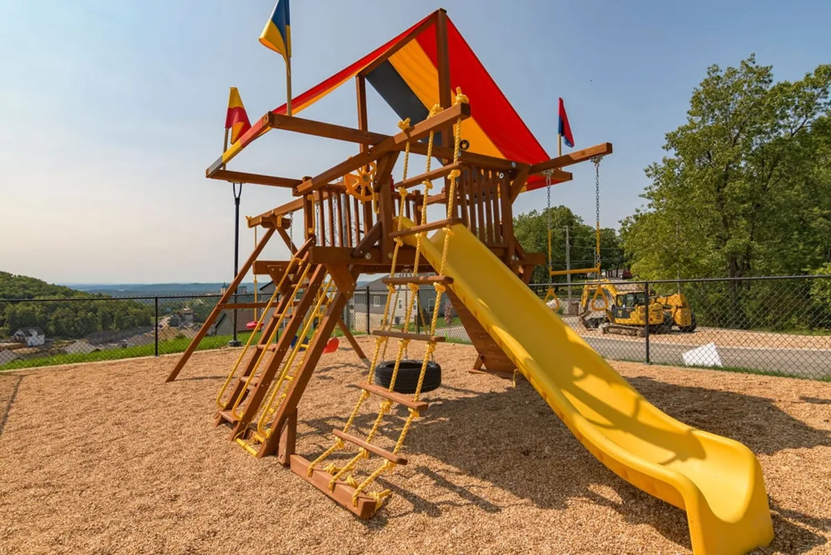 A colorful playground structure takes center stage in this exterior image, complete with a bright yellow slide, climbing elements, and festive flags. The joyous mood is underscored by visible children’s play and the surrounding greenery.
