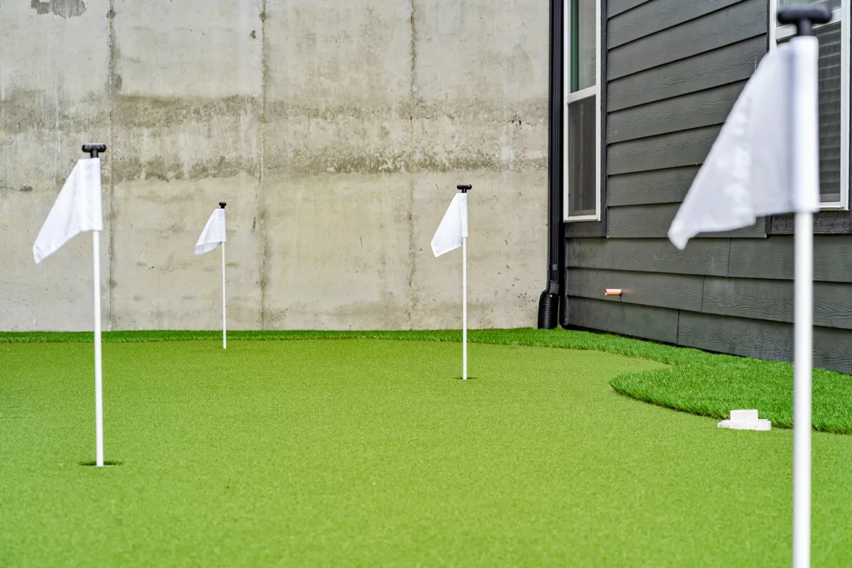 This image showcases a vibrant outdoor putting green surrounded by lush artificial turf and bordered by a concrete wall. White flags mark the holes, suggesting a space designed specifically for putting practice, while the open sky above and well-maintained grounds create an inviting setting for outdoor recreation.