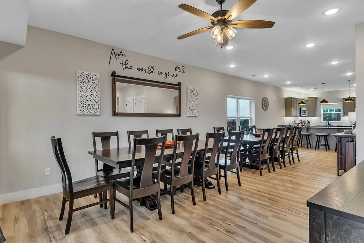 A broad view of a large dining table set for a meal, with decorative wall art and a mirror reflecting the adjoining kitchen. The setting highlights the capacity for family feasts or festive dinner parties, with an ambiance that feels both homey and celebratory.