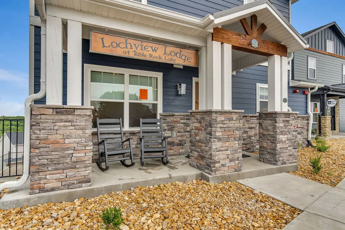 The image showcases the welcoming exterior of Lochview Lodge, featuring a prominent lodge sign, inviting rocking chairs, and stone pillars on a spacious porch. In the background, the lake and lush surroundings hint at the scenic and tranquil setting, perfect for family arrivals and departures.
