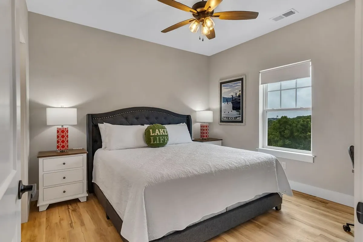 A tranquil king bedroom featuring a bed with decorative pillows, nightstands with lamps, tasteful lake-themed wall art, and a window offering natural light and an inviting view. The calming tones and plush bedding create a relaxing atmosphere for restful nights.