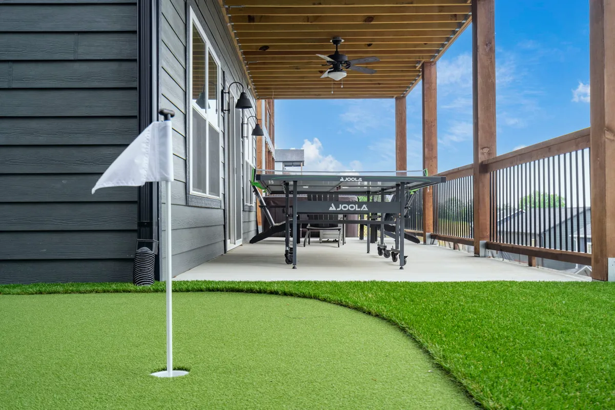 A spacious covered outdoor area featuring a putting green, a ping pong table, sturdy wooden beams, and an outdoor ceiling fan. The space feels playful and relaxed, offering opportunities for both golf practice and casual games, with natural light and landscaped surroundings visible.