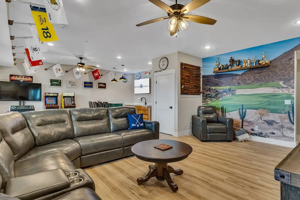 The image showcases a spacious, inviting living area featuring a plush leather sectional sofa, a striking wall mural of a golf course, and sports memorabilia accents. The room is designed for relaxation and entertainment, with ample space for guests to gather, play games, or watch TV together around a central wooden table.
