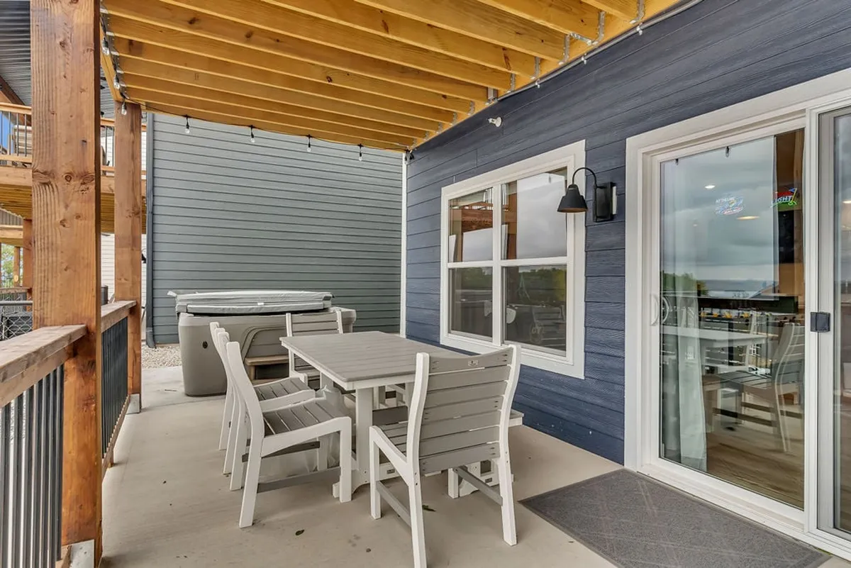 A cozy outdoor patio with a dining table and a luxurious hot tub, all under a wooden-beamed cover. The space is set up for both social gatherings and private relaxation, with comfortable seating and warm outdoor lighting enhancing the inviting mood.