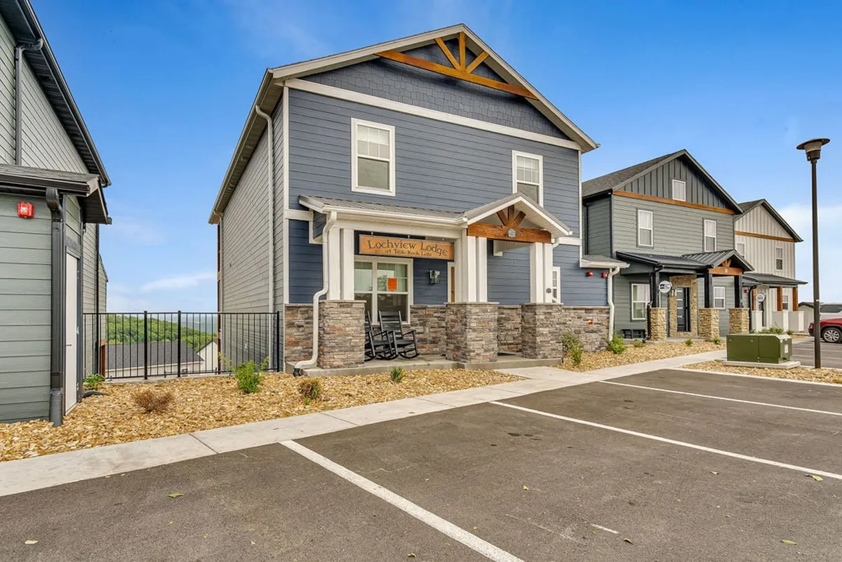 A welcoming exterior view of Lochview Lodge, featuring a stone facade with warm wood accents and a clear sign. The inviting porch area offers space for guests to gather and soak in the setting, with hints of the natural landscape beyond.